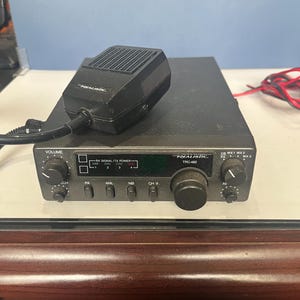 Peut inclure: Une radio CB Realistic TRC-482 noire avec un microphone assorti. La radio comporte plusieurs boutons et molettes, notamment pour le volume, la sélection des canaux et PA. Le microphone est posé sur le dessus de la radio.