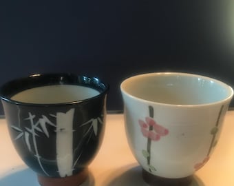 Puede incluir: Dos pequeñas tazas de té de cerámica con diseños diferentes. Una taza es negra con diseños de bambú blancos, la otra es blanca con flores rosas y hojas verdes.
