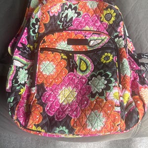 Puede incluir: Una mochila acolchada con un vibrante estampado floral en tonos rosa, naranja, amarillo y verde sobre un fondo negro. La mochila tiene múltiples compartimentos y un bolsillo frontal.