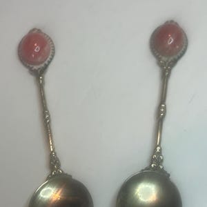 May include: Two silver spoons with pink and white shell-shaped handles. The spoons have a rounded bowl shape.