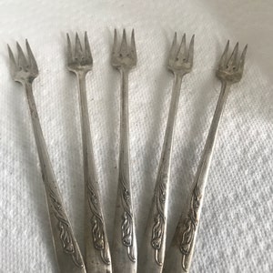 Può includere: Set di cinque forchette da cocktail argentate con decorazioni floreali ornate sui manici.