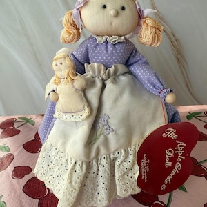 May include: A purple and white fabric doll with blonde hair and a white dress. The doll is holding a smaller doll in her arms. The doll is sitting on a pink and red background with a cherry pattern. The doll has a tag that says "The Apple Cheeks Doll" on it.