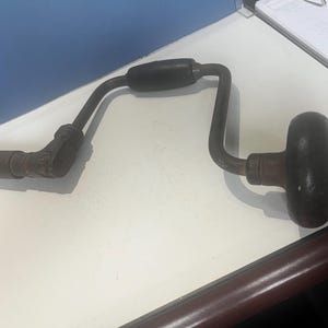 Puede incluir: Un taladro manual antiguo con un mango de madera oscura y un marco de metal curvado. El taladro tiene un diseño clásico, con un mango redondeado en un extremo y un portabrocas para sujetar las brocas en el otro. El metal muestra signos de edad.