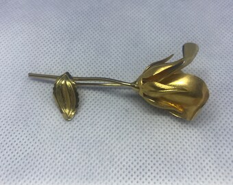 Könnte beinhalten: Eine goldfarbene Rosenbrosche mit einem einzelnen Blatt. Die Rose ist stilisiert und hat eine strukturierte Oberfläche.