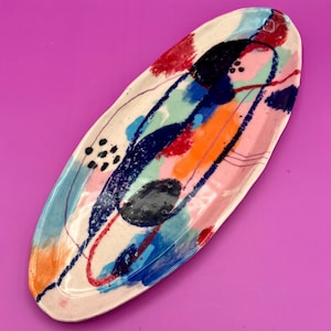 May include: An oval ceramic serving dish with a white background and abstract colorful designs in blue, red, orange, pink, and black.