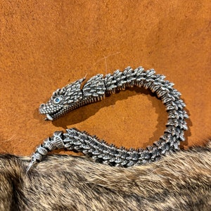 May include: A silver bracelet with a dragon design. The dragon's head is detailed with blue eyes and scales. The bracelet is made of interlocking pieces that form a flexible chain.