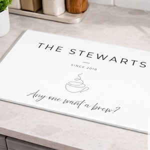 May include: White rectangular kitchen cutting board with the text "THE STEWARTS SINCE 2018" and a coffee cup graphic. Below the graphic is the text "Any one want a brew?" The board is on a light grey countertop.