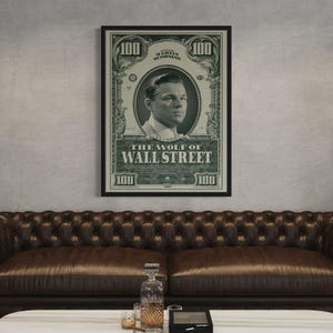 May include: Framed poster of Leonardo DiCaprio from "The Wolf of Wall Street", designed as a $100 bill. The black-framed poster hangs above a brown leather sofa. The poster's text reads "The Wolf of Wall Street".