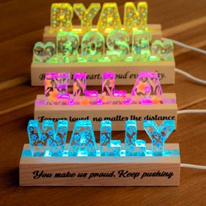 May include: Three custom light-up name signs made of resin with embedded floral patterns, set on rectangular wooden bases. The signs display the names RYAN, ROSE, ELLA, and WALLY in various colors, with inspirational text printed on the wooden bases.