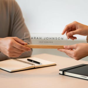 May include: A clear acrylic desk nameplate with the name "SARAH JOHNSON" and the title "CASE MANAGER" printed in black. The nameplate is set on a light-colored wooden base. The nameplate is being held by two people.