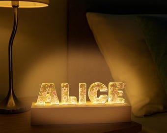 May include: A light-up sign displaying the name ALICE in block letters. Each letter is made of a clear material filled with a colorful floral pattern of pink, white, and yellow flowers. The letters are mounted on a rectangular wooden base.