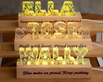 May include: Three illuminated wooden name signs with resin letters. The top sign spells ELLA with yellow daffodils. The middle sign spells ROSE with heart icons and floral resin. The bottom sign spells WALLY with blue and yellow flowers.