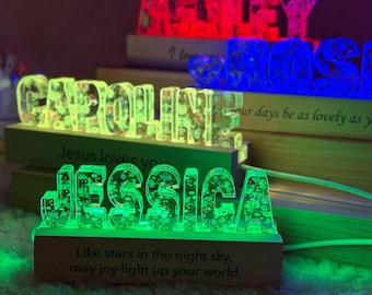 May include: A series of custom name signs made of clear resin with embedded floral patterns, illuminated by LED bases. The front sign displays the name JESSICA in green light, with the text: Like stars in the night sky, may joy light up your world.