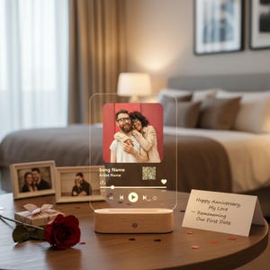 May include: A clear acrylic photo display with a wooden base, featuring a couple's photo and music player interface. The display includes the text "Song Name" and "Artist Name". A red rose and a gift box are on the table.