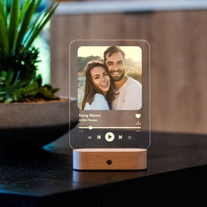 May include: A clear acrylic music player with a photo of a couple on a beach. The player has a heart icon, a download icon, and a play button. The text "Song Name" and "Artist Name" are displayed on the screen.
