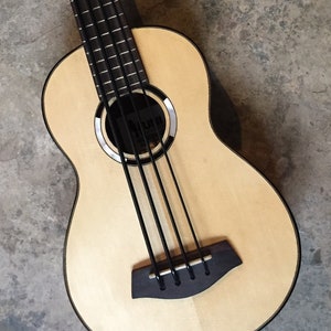 May include: A wooden ukulele bass with four strings and a natural wood finish. The bass has a black pickguard and black tuning pegs.