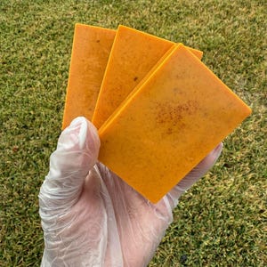 May include: Three rectangular bars of orange soap are held in a gloved hand against a backdrop of green grass. The soap has a textured surface and a speckled appearance, suggesting natural ingredients.