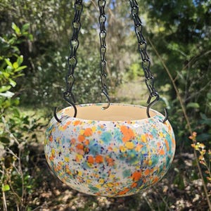 May include: A hanging ceramic planter with a speckled pattern in orange, blue, and yellow. The planter is suspended by a black chain. The interior of the planter is a cream color.
