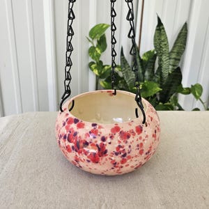 May include: A round ceramic hanging planter with a glossy finish, featuring a light pink base color with speckled patterns in shades of red, purple, and white. The bowl is suspended by three black metal chains attached to the rim.