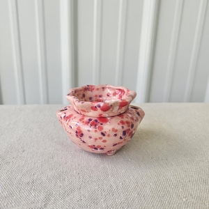 May include: A small, round ceramic vessel with a scalloped rim and a speckled glaze in shades of light pink, dark red, and purple. The bowl-shaped base features two small openings on the sides, creating a distinct two-tiered appearance.