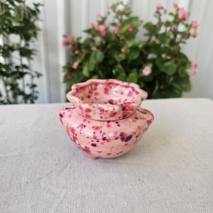 May include: A small, round ceramic bowl with a scalloped rim and a speckled glaze in shades of light pink, dark pink, and purple. The bowl features a wide, rounded base and a distinct upper section, creating a layered appearance with a glossy finish.