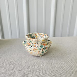 May include: A small ceramic bowl with a scalloped rim and two side handles, featuring a speckled pattern of orange, teal, and yellow dots on a cream-colored base. The bowl has a rounded body and sits on small feet, showcasing a hand-painted finish.