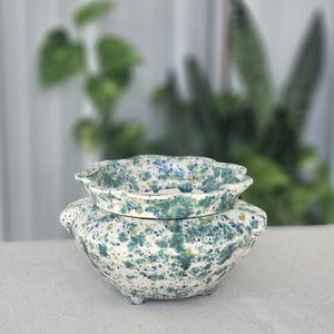 May include: A ceramic planter with a scalloped rim and a rounded body, featuring a speckled glaze in shades of teal, blue, and cream. The vessel is supported by small feet and has a wide opening, showcasing a consistent mottled pattern.