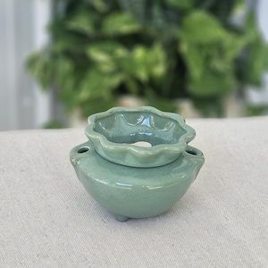 May include: A sage green ceramic oil burner featuring a rounded base with three small feet and a scalloped, flower-shaped top dish. The main body includes two small circular openings on opposite sides for ventilation and heat.
