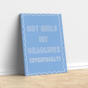 Puede incluir: Un lienzo azul claro con texto blanco que dice "HOT GIRLS HIT DEADLINES (EVENTUALLY)". La impresión tiene un borde festoneado y está apoyada contra una pared blanca sobre un suelo de madera.