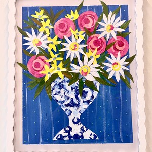 May include: A framed floral art piece featuring a vase filled with paper flowers. The flowers include pink roses, white daisies, and yellow star-shaped blooms. The vase has a blue and white pattern, set against a blue background with white dots. The frame is white.