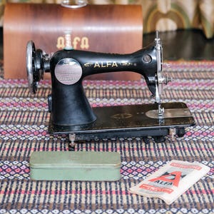 Könnte beinhalten: Eine schwarze Vintage-Nähmaschine von Alfa mit silbernen Details. Sie steht auf einem gemusterten Teppich, im Hintergrund befindet sich ein brauner Lederkoffer. Eine grüne Metallbox und ein Nadelpaket sind ebenfalls vorhanden.