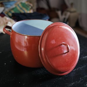 Può includere: Una pentola da cucina in smalto rosso con coperchio abbinato. La pentola ha un manico su un lato e un interno azzurro chiaro. Il coperchio è leggermente socchiuso, rivelando l'interno della pentola. La pentola è su una superficie nera.