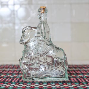 Puede incluir: Una botella de vidrio transparente con forma de persona montada a caballo, con tapón de corcho. La botella tiene detalles intrincados y se encuentra sobre una superficie estampada. La botella mide aproximadamente 20 cm de altura.