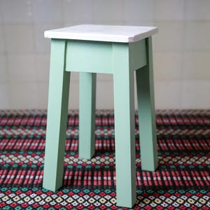 Puede incluir: Un pequeño taburete cuadrado de madera pintado en un suave verde menta con una parte superior blanca. El taburete tiene cuatro patas rectas y está sobre una alfombra estampada. El taburete mide aproximadamente 30 cm de alto.
