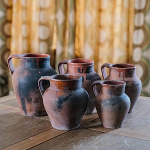 Pode incluir: Um conjunto de seis jarras rústicas de cerâmica com alças. As jarras, de tamanhos variados, apresentam uma mistura de tons castanhos, pretos e avermelhados. Estão dispostas sobre uma superfície de madeira, sugerindo um tema vintage ou artesanal.