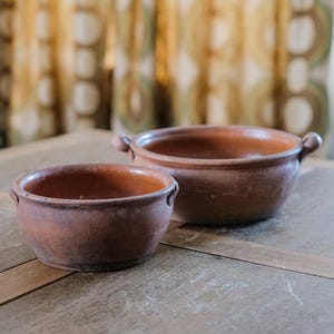 Può includere: Due vasi in terracotta con manici, uno più piccolo dell'altro, su un tavolo di legno.