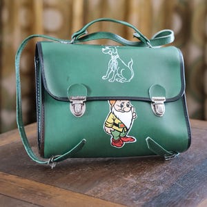 Könnte beinhalten: Grüne Umhängetasche mit oberem Griff und verstellbarem Schultergurt. Die Tasche zeigt eine weiße Umrisszeichnung eines Hundes und eines Cartoon-Zwergs. Die Tasche hat eine schwarze Umrandung und silberne Verschlüsse.