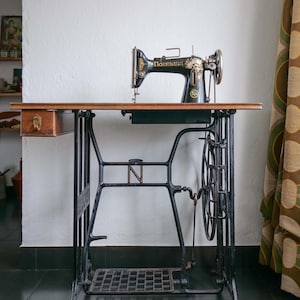 Puede incluir: Una antigua máquina de coser Naumann negra y dorada se encuentra sobre una mesa de madera. La mesa tiene una base de metal negro con un pedal. Un pequeño cajón de madera está adjunto a la mesa.