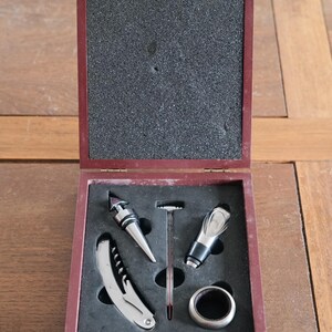 Pode incluir: Um conjunto de ferramentas para vinho em madeira com exterior bordô. O conjunto inclui um saca-rolhas, uma rolha, um bico, um termómetro e um anel para garrafa. As ferramentas estão num interior de espuma preta.