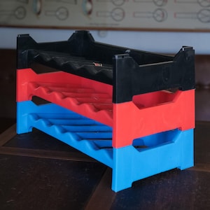 Op de afbeelding: Een wijnrek met drie niveaus van zwart, rood en blauw plastic. Het rek is ontworpen om meerdere flessen wijn te bevatten. Het rek staat op een donkerbruin houten oppervlak.
