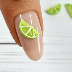 May include: A gold ring with a circular face featuring the initials "N" and "E" with a heart. The image also shows a fingernail with a lime slice design and two additional lime slice decorations.