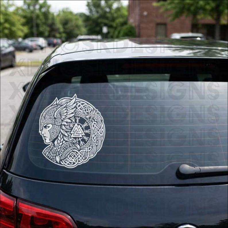 Op de afbeelding: Een witte vinyl sticker van een Viking-krijgervrouw met ingewikkeld knoopwerk en een Vegv&iacute;sir-symbool, aangebracht op de achterruit van een zwarte auto. Het ontwerp toont een profiel van de krijger met een gevleugelde helm.