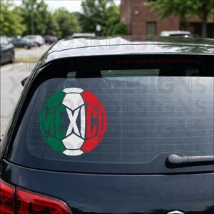 May include: A circular decal applied to the rear window of a black vehicle. The decal features a soccer ball design colored in green, white, and red, representing the Mexican flag, with the word MEXICO integrated into the ball's pattern.