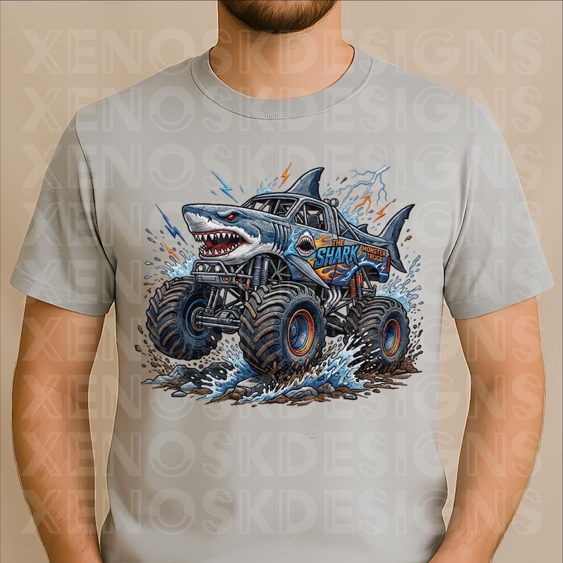 May include: A light grey t-shirt with a shark-themed monster truck design. The truck is blue and white, with the text "The Shark Monster Truck". The image includes water splashes and lightning bolts.
