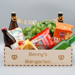 Puede incluir: Una caja de madera con las palabras "Benny's Biergarten" grabadas en la parte delantera. La caja est&aacute; llena de varios bocadillos y bebidas, incluyendo pretzels, patatas fritas y cerveza.