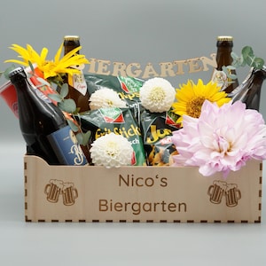 Puede incluir: Una cesta de regalo de madera llena de cerveza, patatas fritas y flores. La cesta est&aacute; decorada con las palabras "Nico's Biergarten".