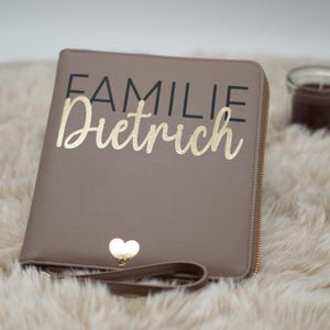 Könnte beinhalten: Ein brauner Lederordner mit Reißverschluss, einem goldenen Herz und dem Text "FAMILIE Dietrich" in schwarzer und goldener Schrift.