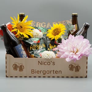 Puede incluir: Una caja de madera llena de cerveza, patatas fritas y flores. La caja est&aacute; grabada con "Nico's Biergarten".