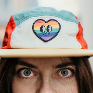 Puede incluir: Una gorra de cinco paneles con un panel frontal color crema y un parche bordado central de un corazón con rayas de arcoíris, ojos de dibujos animados y una sonrisa. La gorra tiene paneles laterales en color verde azulado, detalles rojos y una visera plana de color tostado claro.
