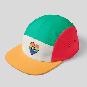 May include: A multi-colored 5-panel cap with a rainbow heart design. The cap is green, white, and red. The heart has a smiling face and is outlined in black.
